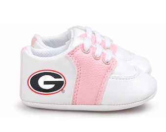 May include: White and pink baby sneakers with white laces. The sneakers feature a pink stripe down the center and a black and red logo on the side. The logo is a black letter "G" inside a red and black oval.