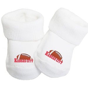 May include: White baby booties with red text that says "Kansas City" and a brown football graphic.