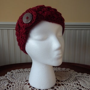 Puede incluir: Una diadema de punto roja con un botón plateado grande. La diadema está en una cabeza de maniquí blanca.
