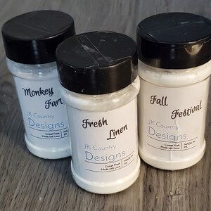 May include: Three glass jars with white labels and black lids. The labels have the text "JK Country Designs" and the names of the scents: "Monkey Fart", "Fresh Linen", and "Fall Festival".
