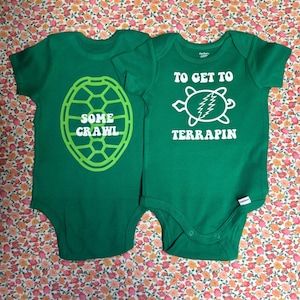 May include: Two green baby onesies with turtle-themed designs. One says "SOME CRAWL" inside a turtle shell outline. The other says "TO GET TO TERRAPIN" with a turtle graphic. Both are on a floral background.