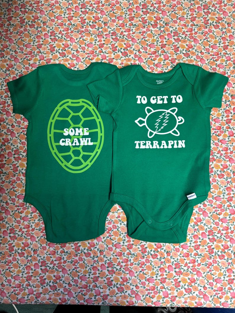 May include: Two green baby onesies with turtle-themed designs. One says "SOME CRAWL" inside a turtle shell outline. The other says "TO GET TO TERRAPIN" with a turtle graphic. Both are on a floral background.