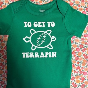 May include: Green baby bodysuit with the text "TO GET TO TERRAPIN" in white. The design includes a white turtle graphic with a lightning bolt. The bodysuit is made by Gerber and has short sleeves.