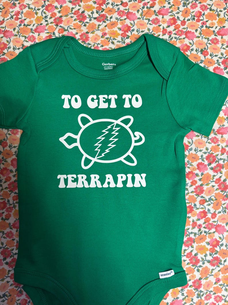 May include: Green baby bodysuit with the text "TO GET TO TERRAPIN" in white. The design includes a white turtle graphic with a lightning bolt. The bodysuit is made by Gerber and has short sleeves.