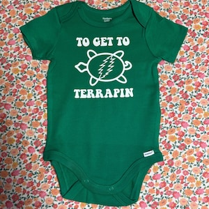 May include: Green baby onesie with the text "TO GET TO TERRAPIN" in white. A white turtle graphic with a lightning bolt design is centered below the text. The onesie is made of soft material and has short sleeves.
