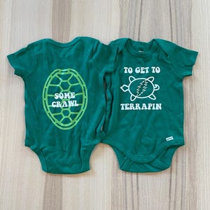 May include: Two green baby onesies with turtle-themed designs. One features a turtle shell graphic with the words "SOME CRAWL." The other has a turtle graphic with the text "TO GET TO TERRAPIN." Both are short-sleeved.