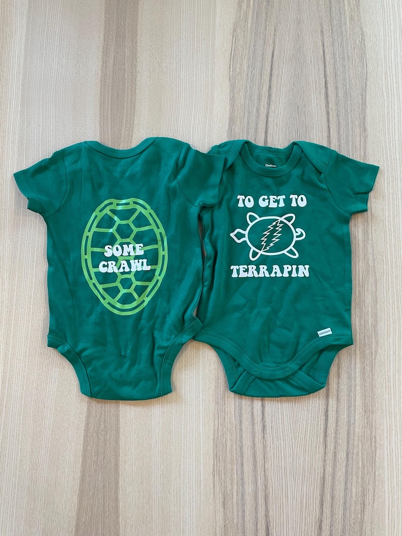 May include: Two green baby onesies with turtle-themed designs. One features a turtle shell graphic with the words "SOME CRAWL." The other has a turtle graphic with the text "TO GET TO TERRAPIN." Both are short-sleeved.