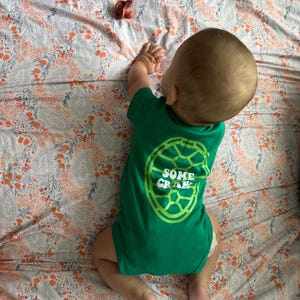 May include: A baby wearing a green onesie with the text "SOME CRAWL" in a lime green turtle shell design. The baby is lying on a floral patterned blanket with orange and blue accents. A pacifier and a toy are also on the blanket.