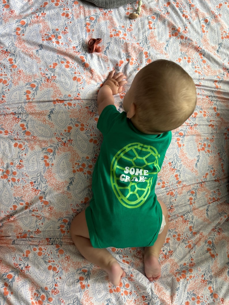 May include: A baby wearing a green onesie with the text "SOME CRAWL" in a lime green turtle shell design. The baby is lying on a floral patterned blanket with orange and blue accents. A pacifier and a toy are also on the blanket.