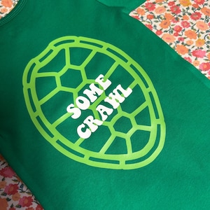 May include: Green baby bodysuit with a lime green turtle shell design and the words "SOME CRAWL" in white. The bodysuit is made of soft fabric and is likely for a baby or toddler.