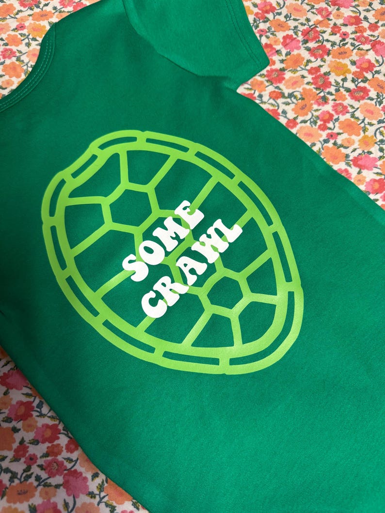 May include: Green baby bodysuit with a lime green turtle shell design and the words "SOME CRAWL" in white. The bodysuit is made of soft fabric and is likely for a baby or toddler.