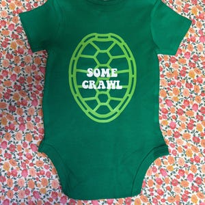 May include: A green baby bodysuit with short sleeves. The bodysuit features a white and green turtle shell design on the front, with the words "SOME CRAWL" in white letters inside the shell.
