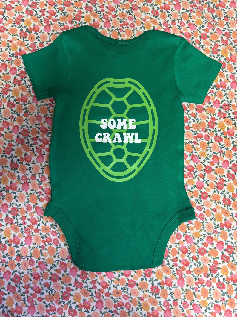May include: A green baby bodysuit with short sleeves. The bodysuit features a white and green turtle shell design on the front, with the words "SOME CRAWL" in white letters inside the shell.