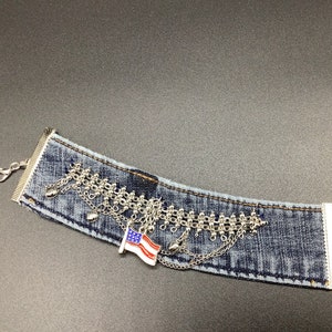 Puede incluir: Un brazalete de mezclilla con detalles de cadena plateada y un pequeño dije de bandera estadounidense.