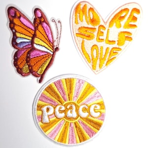 May include: Three embroidered iron-on patches with a yellow and pink color scheme. The patches feature a butterfly, a heart with the text "More Self Love", and a sunburst with the text "peace".