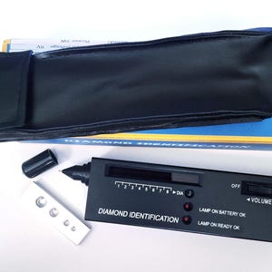 May include: A black diamond identification tool with a silver metal testing plate and a black carrying case. The tool has a digital display and reads "DIAMOND IDENTIFICATION". It has an on/off switch and volume control.