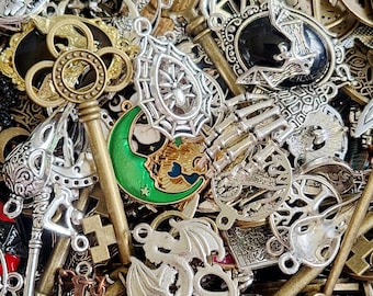 May include: A collection of metal charms and pendants featuring various designs including a large skeleton key, a spider web, a bat, a green crescent moon, and a dragon. The assortment includes silver and gold-toned metal pieces.