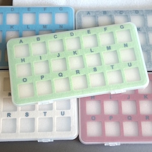 May include: A collection of five rectangular plastic storage cases, each featuring a grid of twenty-one square compartments. The cases are colored blue, light green, white, gray, and pink, with letters A through U printed above each slot.