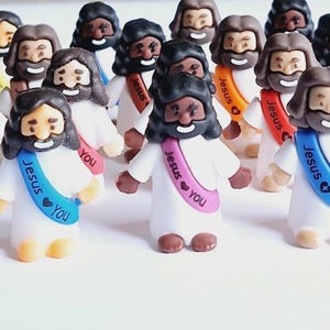 May include: A collection of small figurines depicting Jesus, each with a different skin tone and hair color. They wear white robes and sashes with the words "Jesus ❤️ You." The figurines have smiling faces and are arranged on a white surface.