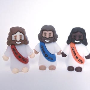 May include: Three small figurines of Jesus in white robes, each with a different skin tone and hair color. Each figure has a sash that reads "Jesus You". The figurines are holding hands and have smiling faces.