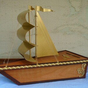 A wooden model sailboat with a gold painted hull and sails. The sailboat has a rope railing and decorative gold accents. The sailboat is sitting on a blue surface.
