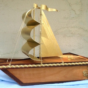 May include: A wooden model sailboat with gold sails and a brown wooden base. The sailboat has a rope railing around the deck and gold accents on the base.