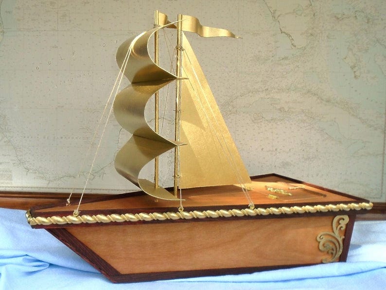 May include: A wooden model sailboat with gold sails and a brown wooden base. The sailboat has a rope railing around the deck and gold accents on the base.