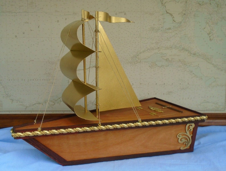 A wooden model sailboat with a gold painted hull and sails. The sailboat has a rope railing and decorative gold accents. The sailboat is sitting on a blue surface.