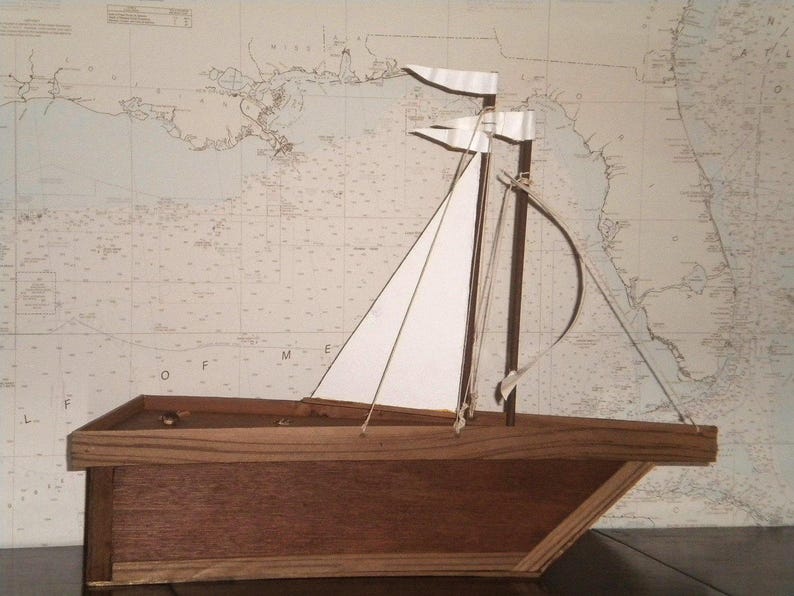 A wooden model sailboat with white sails. The boat is sitting on a wooden base. The background is a nautical chart with a brown and white color scheme.
