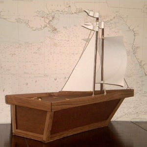 A small wooden model sailboat with white sails. The sailboat is sitting on a wooden surface. The sailboat has a brown wooden hull and a white sail. The sail is partially unfurled. The sailboat is in front of a white background with a map on it.