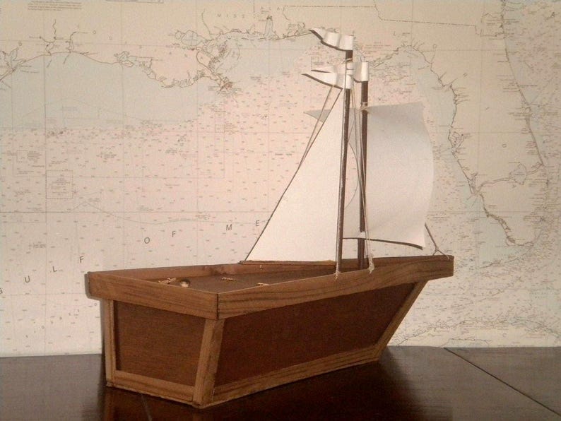 A small wooden model sailboat with white sails. The sailboat is sitting on a wooden surface. The sailboat has a brown wooden hull and a white sail. The sail is partially unfurled. The sailboat is in front of a white background with a map on it.