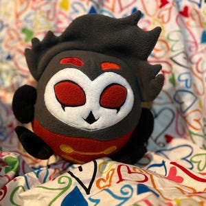 May include: A plush toy featuring a stylized character with a dark gray head and body, accented with red and white details. The character has large red eyes, a white face, and a small black nose. The toy is resting on a colorful heart-patterned fabric.