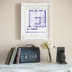 May include: A framed blueprint of a house floor plan in blue ink on white paper. The frame is white with ornate detailing. The blueprint is displayed on a white wall above a white table with a vintage camera, a lunchbox, and a vase of white flowers.