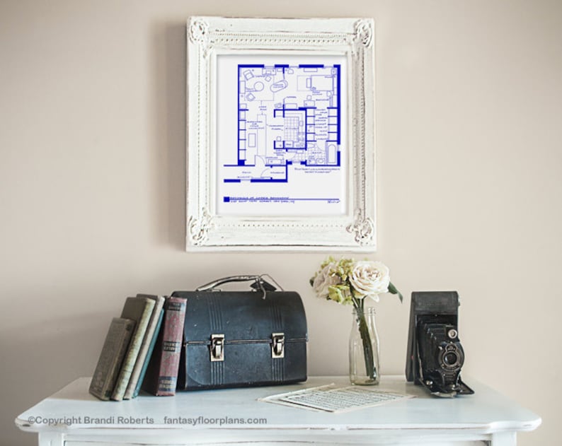 May include: A framed blueprint of a house floor plan in blue ink on white paper. The frame is white with ornate detailing. The blueprint is displayed on a white wall above a white table with a vintage camera, a lunchbox, and a vase of white flowers.