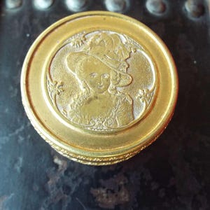 Peut inclure: Une boîte en métal doré avec un motif en relief d'une femme portant un chapeau et une robe. La boîte a une bordure décorative.