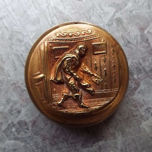 Puede incluir: Un botón dorado con un diseño en relieve de una persona con una túnica caminando a través de una puerta. El botón tiene un borde decorativo.