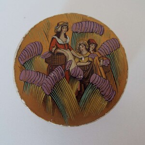May include: A round, gold-coloured tin with a vintage illustration of three women in a field of purple flowers. The women are wearing dresses and carrying baskets. The tin has a decorative border.