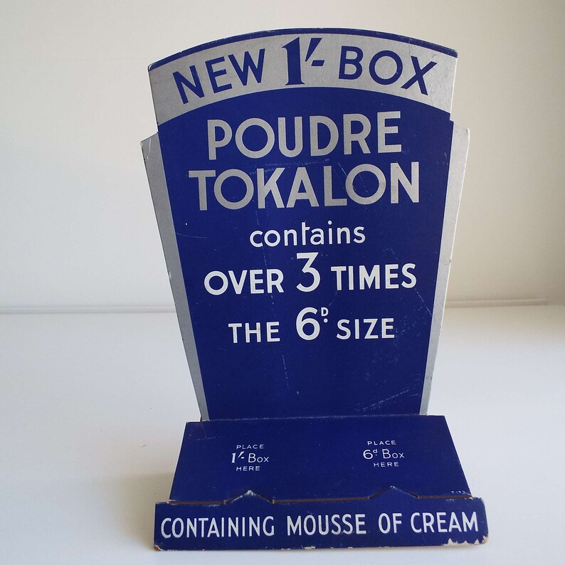 以下が含まれることがあります： 青と銀色の段ボール製のディスプレイスタンドが、「ニュー1/2ボックス プードル トカロン」と宣伝し、「6dサイズの3倍以上」と記載されています。スタンドには、「1/2ボックス」と「6dボックス」のラベルが付けられた2つのボックス用のスロットがあります。スタンドの下部には、「ムースクリーム入り」と記載されています。