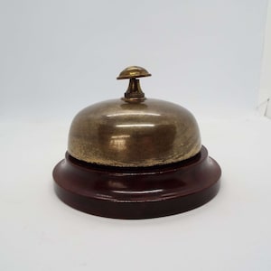 May include: A brass bell with a wooden base. The bell is gold in colour and has a small knob on top. The base is dark brown.