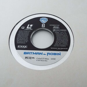 May include: A silver laser disc with a black and white image of Batman and Robin. The disc is labeled "Batman & Robin" and has a PG-13 rating. The disc is also labeled "Extended Play" and "Digitally Processed".