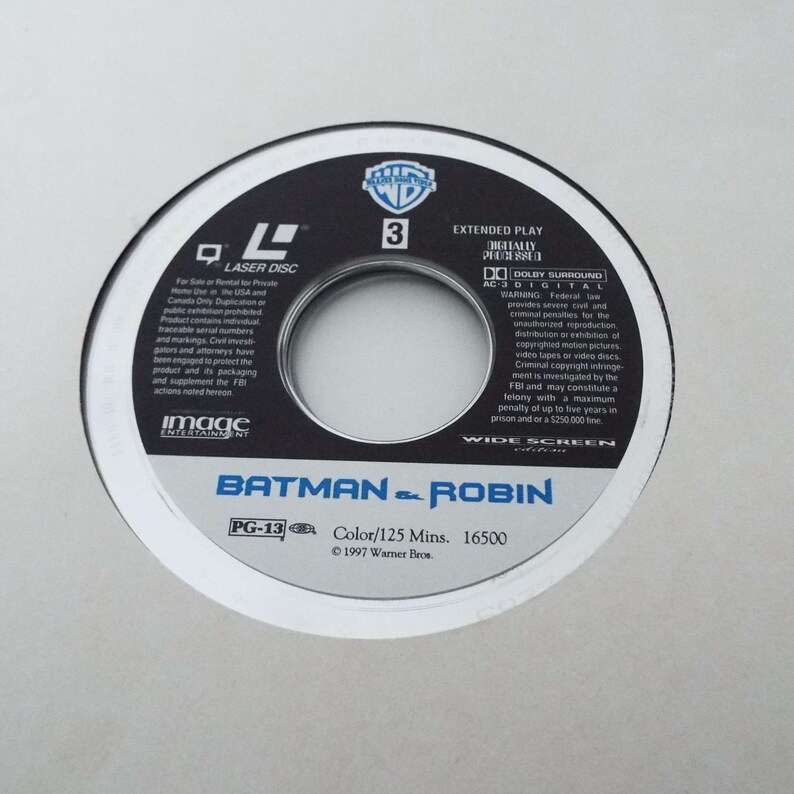 May include: A silver laser disc with a black and white image of Batman and Robin. The disc is labeled "Batman & Robin" and has a PG-13 rating. The disc is also labeled "Extended Play" and "Digitally Processed".