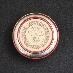 May include: A vintage, round face powder tin with a red and silver rim. The center label reads "SAMPLE LEICHNER Face Powder LONDON" surrounded by a floral wreath. The tin is set against a dark background.