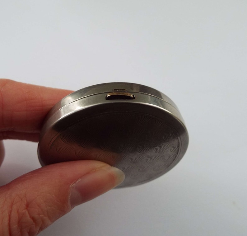 May include: A silver compact with a textured, circular design and a gold latch.