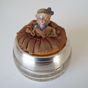May include: A vintage glass jar with a silver lid and a porcelain doll head on top. The doll is wearing a straw hat with a blue bow and a white dress. The jar is filled with a brown fabric and has a ruffled edge.