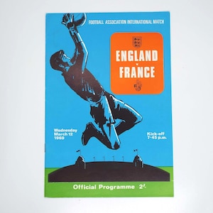 May include: A vintage football match program for England v France on March 12, 1969. The light blue cover features a dark blue illustration of a goalkeeper jumping to catch a ball, an orange rectangular box with white text, and a green base.