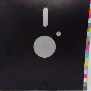 Pode incluir: Uma capa de disco de vinil quadrada preta com um recorte circular central e um recorte retangular menor acima, imitando o design de um disquete. A borda direita exibe uma coluna vertical de pequenos quadrados multicoloridos em vários tons.