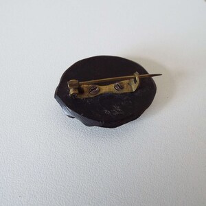 May include: A dark brown wooden brooch with a gold metal pin and clasp. The brooch is oval shaped and has a smooth surface.