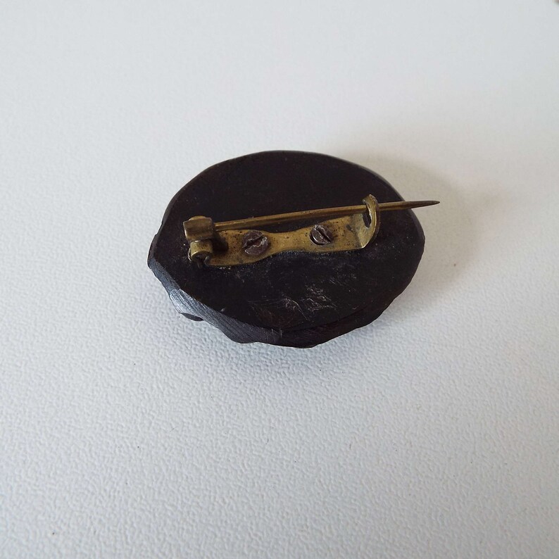 May include: A dark brown wooden brooch with a gold metal pin and clasp. The brooch is oval shaped and has a smooth surface.