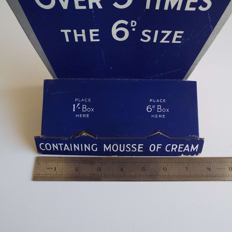 以下が含まれることがあります： 「THE 6 SIZE」と「CONTAINING MOUSSE OF CREAM」の文字が書かれた青と白の段ボール製ディスプレイスタンド。スタンドには、「PLACE 1- Box HERE」と「PLACE 6- Box HERE」というラベルの付いたスロットが2つあります。
