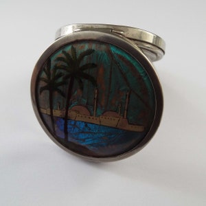 May include: A silver compact with a round, hinged lid. The lid is decorated with a colorful scene of palm trees and buildings reflected in a blue body of water. The scene is made from iridescent butterfly wings.